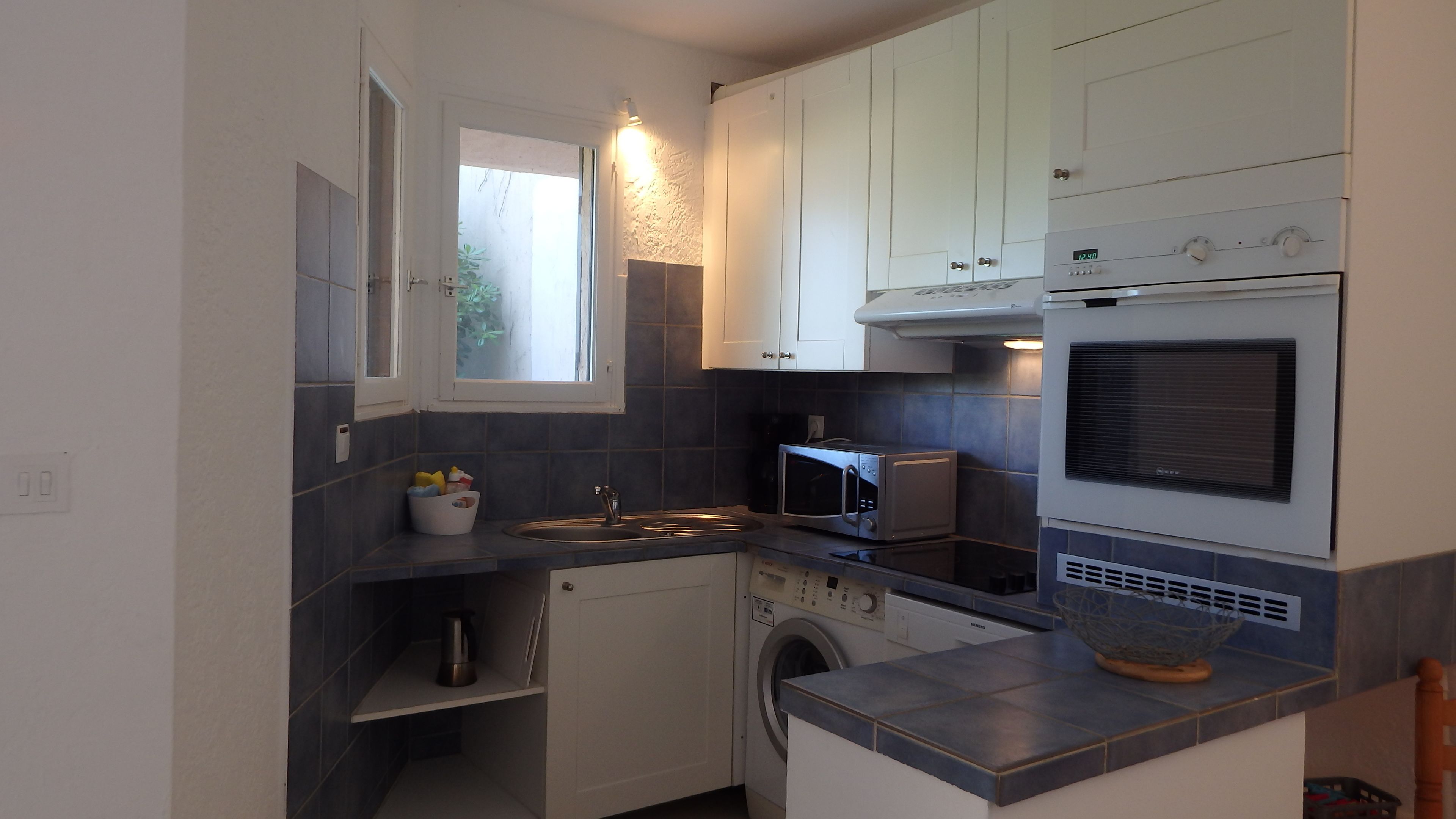 A compact kitchen with white cabinets, a built-in microwave, a washing machine, and a window providing natural light.