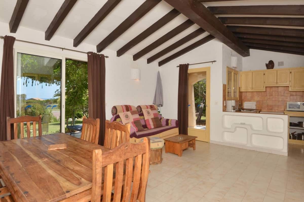 A cozy room with a wooden dining table, a sofa with colorful cushions, a kitchen area with wooden cabinets, and a sliding glass door leading to a garden.
