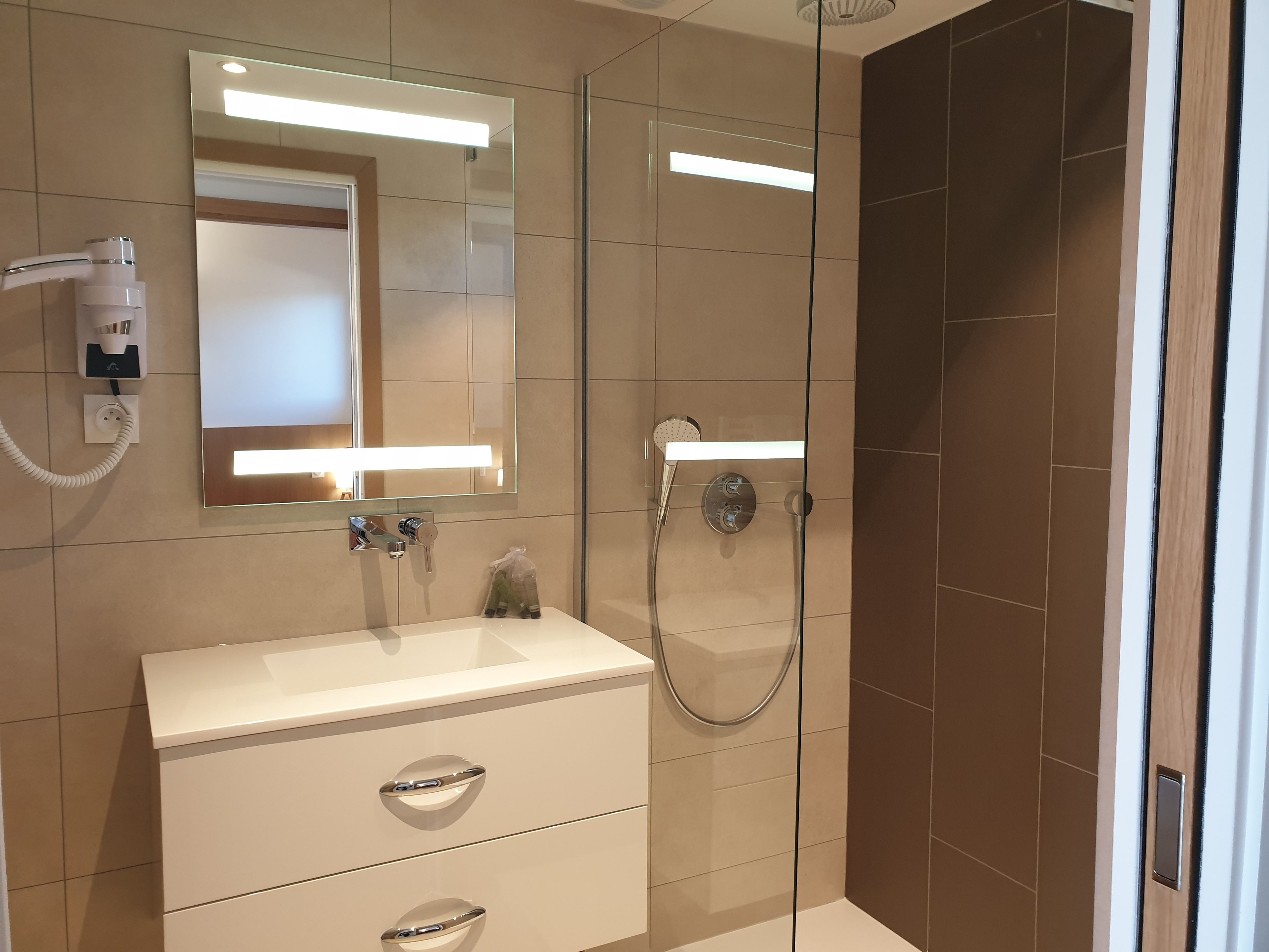 A modern bathroom with a glass-enclosed shower, a white vanity with a mirror, and a hairdryer mounted on the wall.