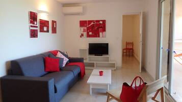 A modern living room with a grey sofa, red cushions, a flat-screen TV, a white coffee table, and a red bag.