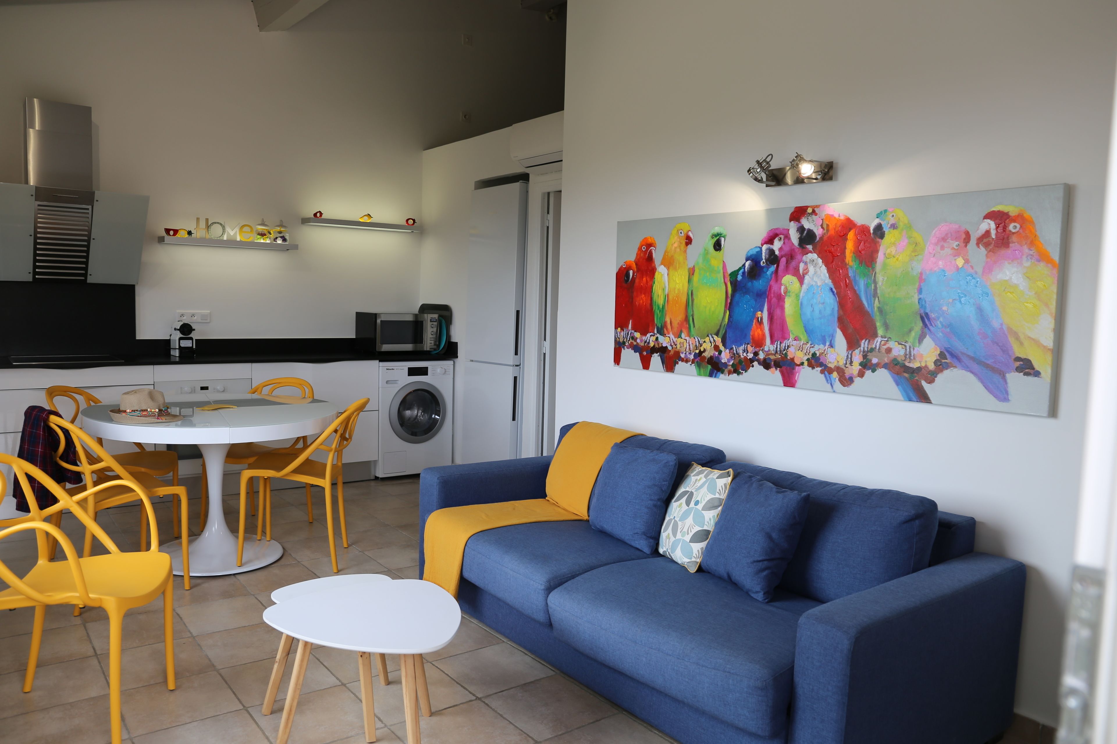 A modern living room with a blue sofa and yellow chairs, a dining table, and a kitchen in the background.