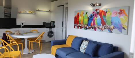 A modern living room with a blue sofa and yellow chairs, a dining table, and a kitchen in the background.