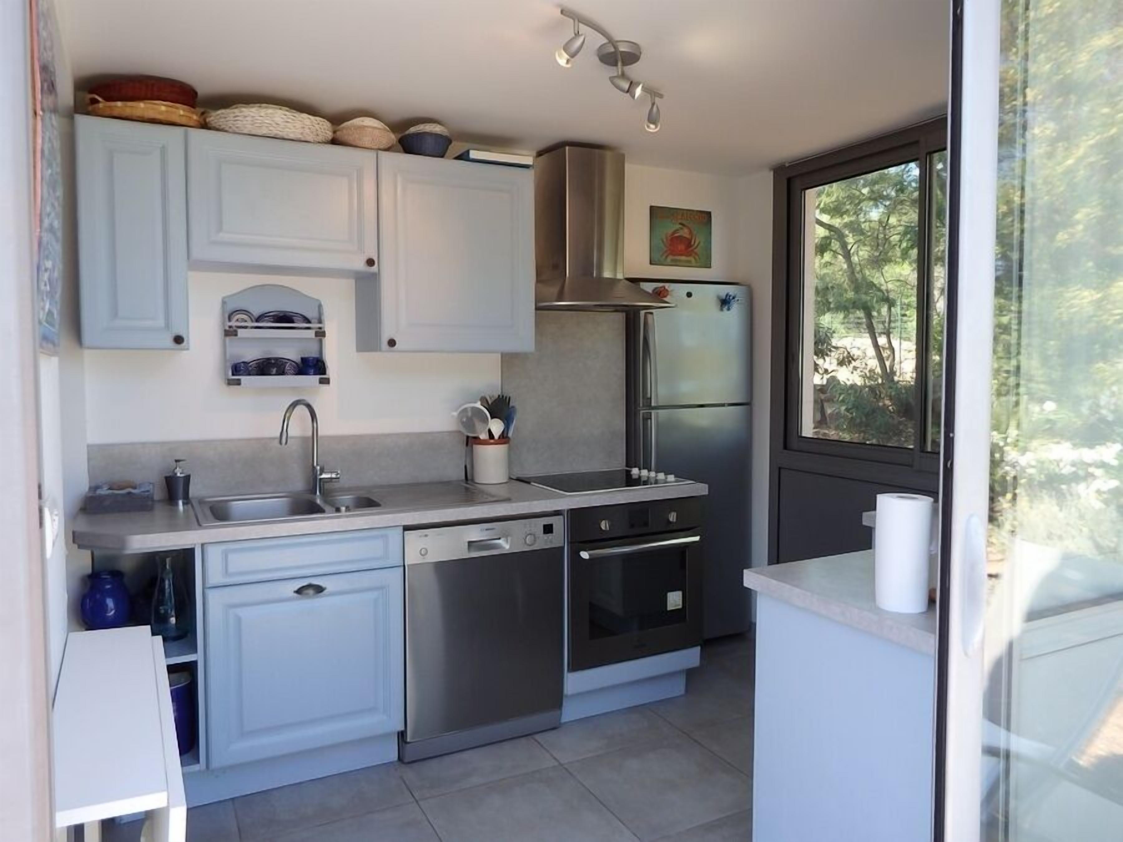 A modern kitchen with light blue cabinets, stainless steel appliances, and a large window offering a view of greenery.