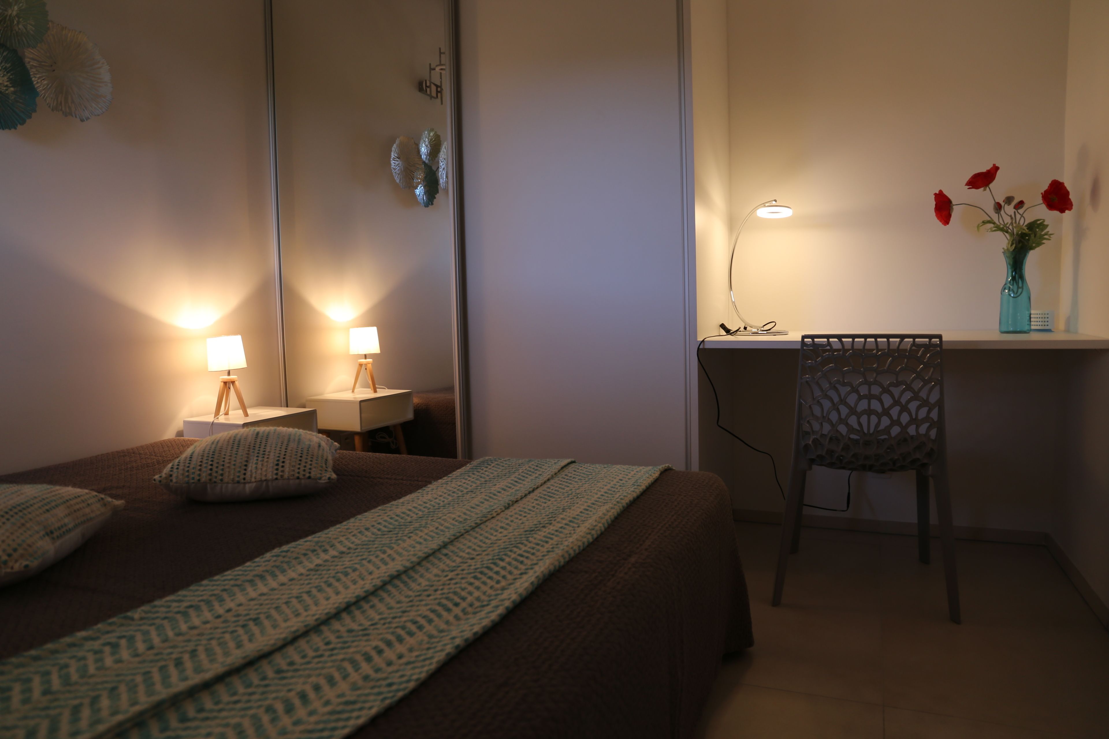 Un dormitorio con cama, lámparas de noche, escritorio, silla y un florero con flores.