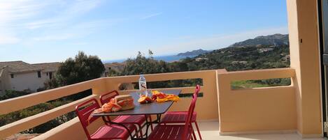 A balcony with a table set for two, red chairs, and a view of the sea and hills.
