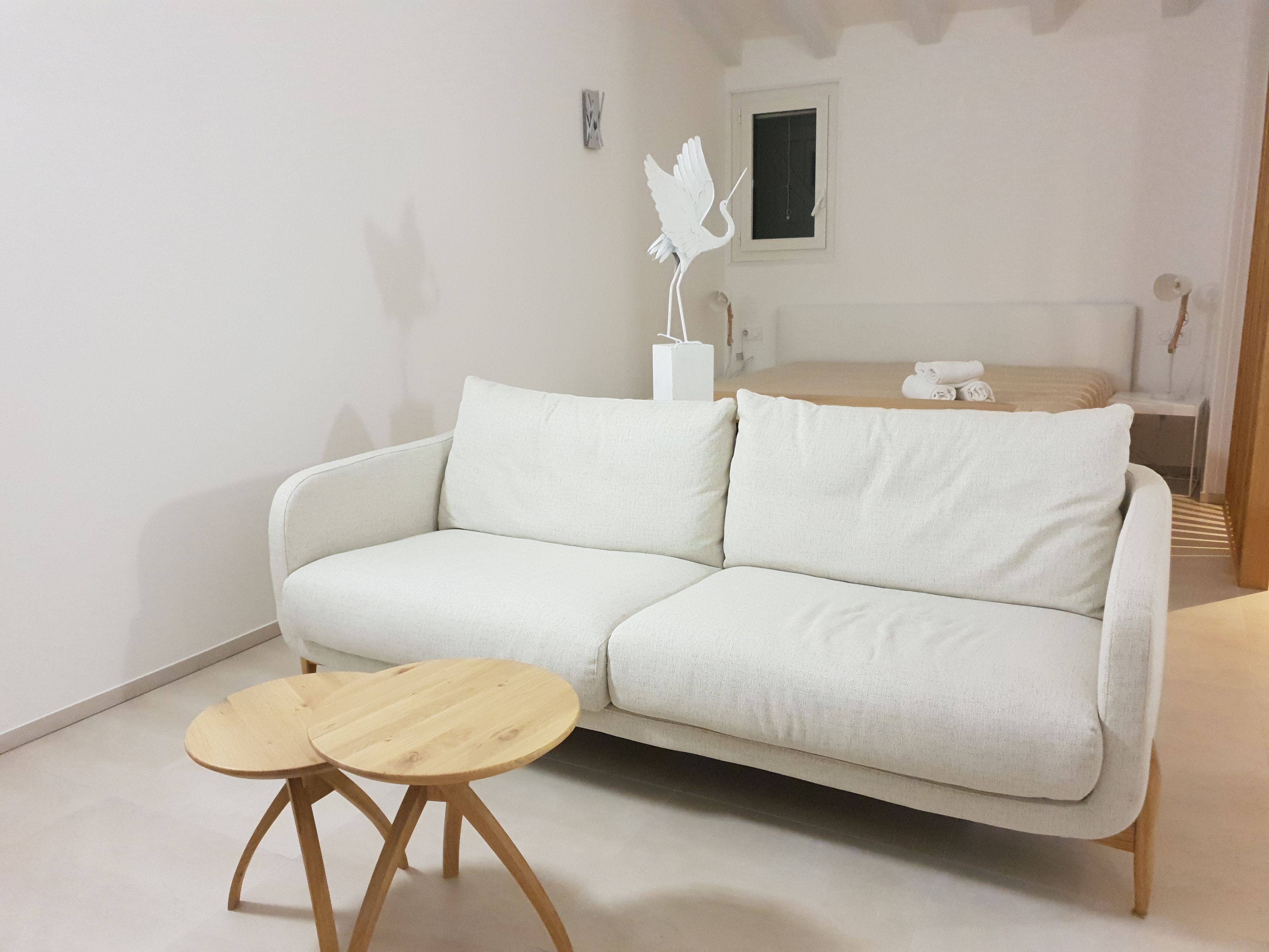 A white sofa with two round wooden tables in a minimalist room with a bed and a window.