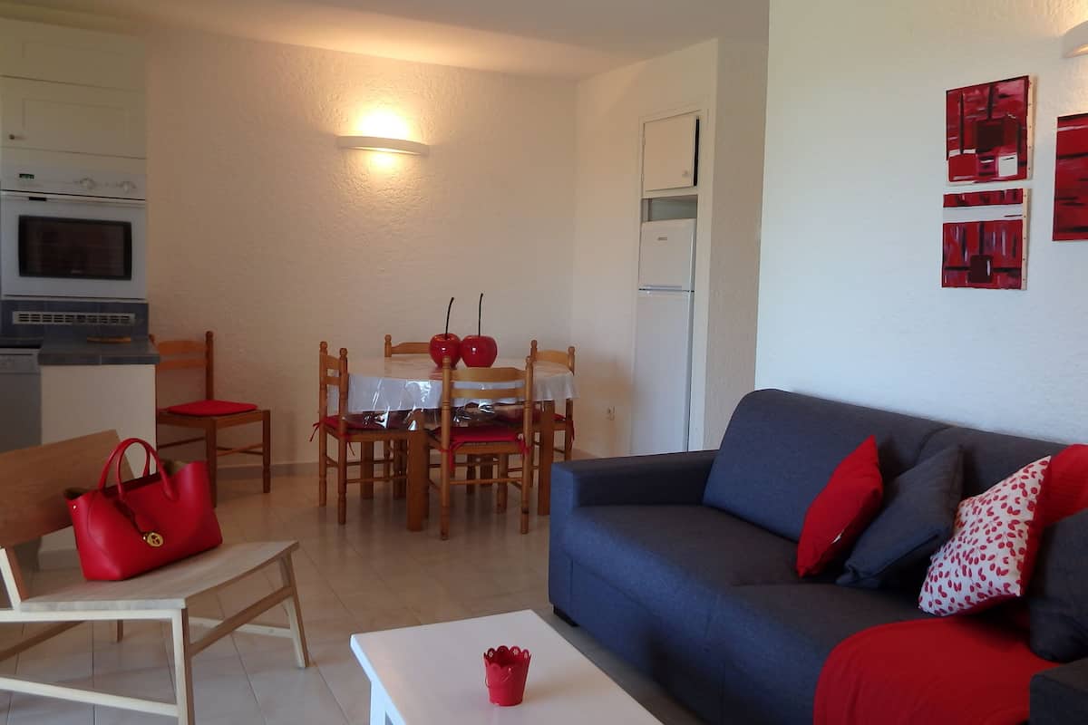 A living room with a sofa, a dining table with chairs, a kitchen area with an oven, and a red handbag on a chair.