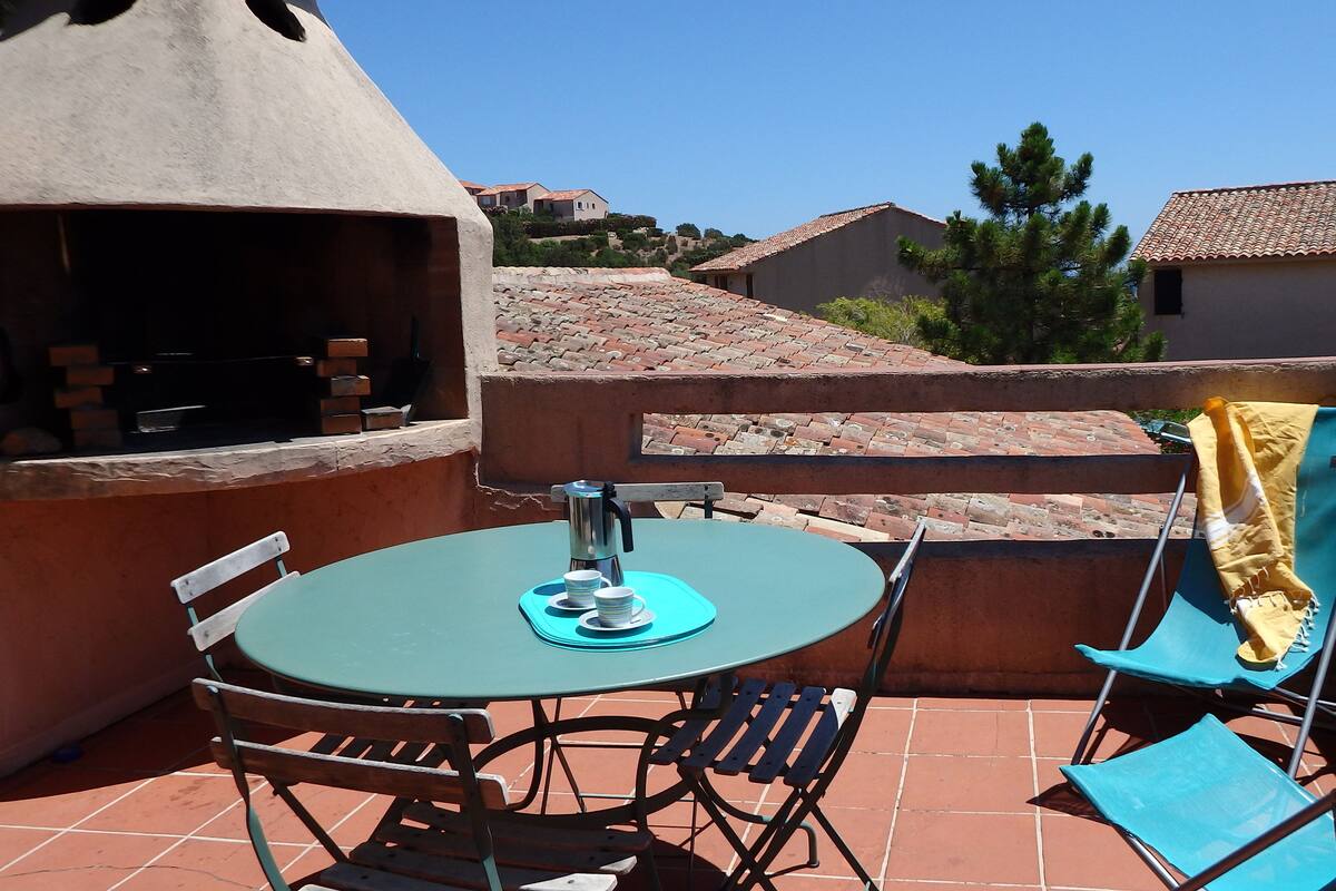 A patio with a round table, chairs, a grill, and a view of neighboring houses.