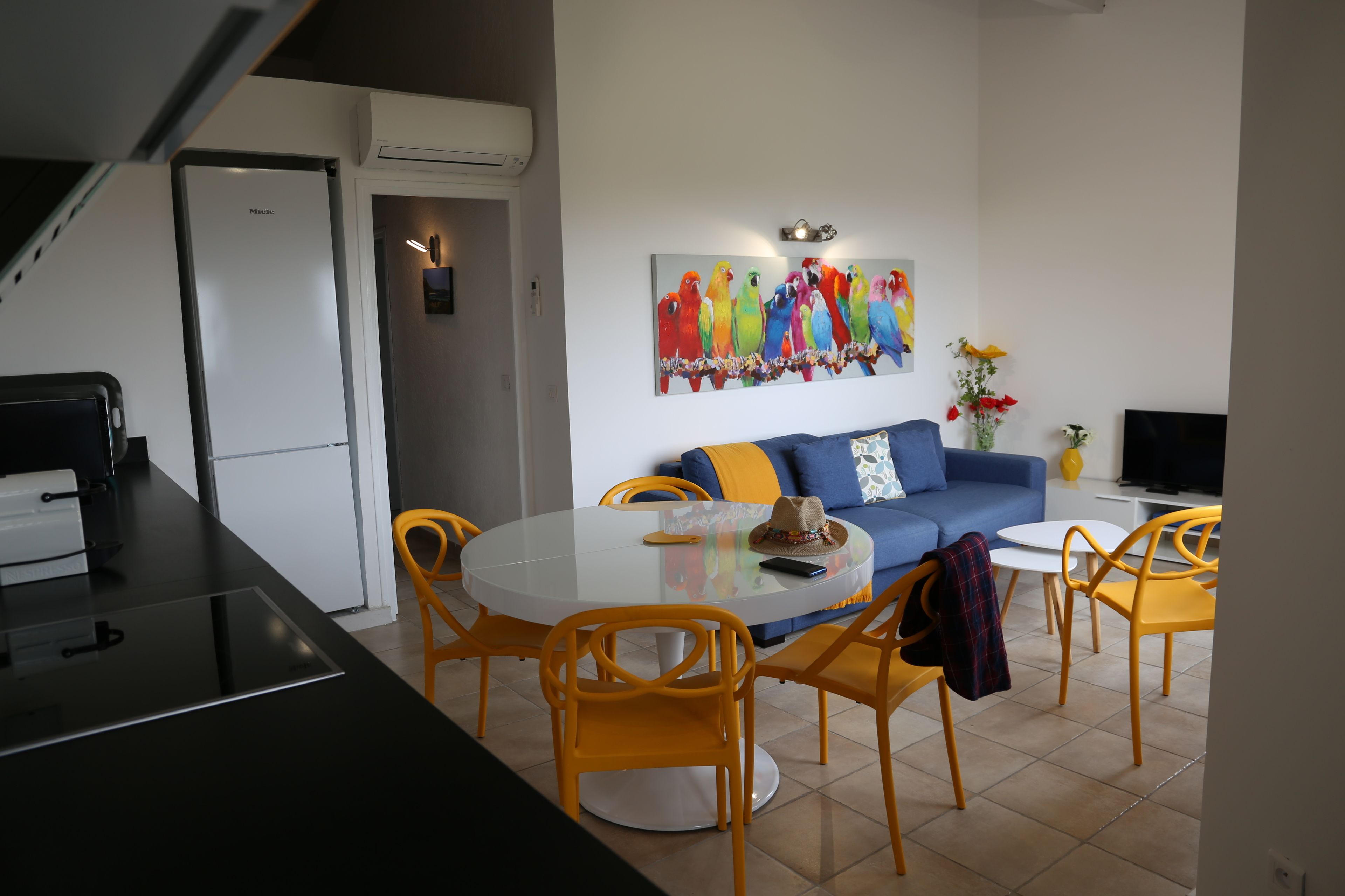 A modern kitchen with a refrigerator, dining table with chairs, and a living area with a sofa and a colorful parrot painting.