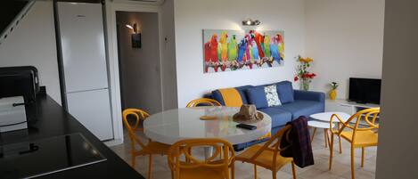 A modern kitchen with a refrigerator, dining table with chairs, and a living area with a sofa and a colorful parrot painting.