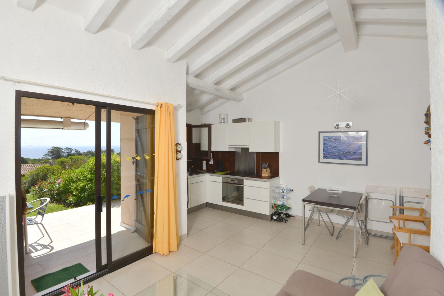 Una cucina moderna con mobili bianchi, una zona pranzo con tavolo e sedie, e una porta scorrevole in vetro che dà su un balcone con vista su spazi verdi e sul mare.