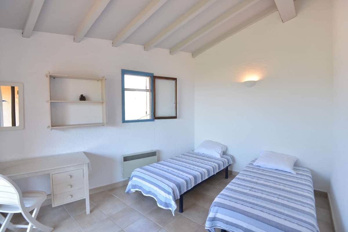 A small, clean room with two single beds, a desk, a chair, and a window with a view.