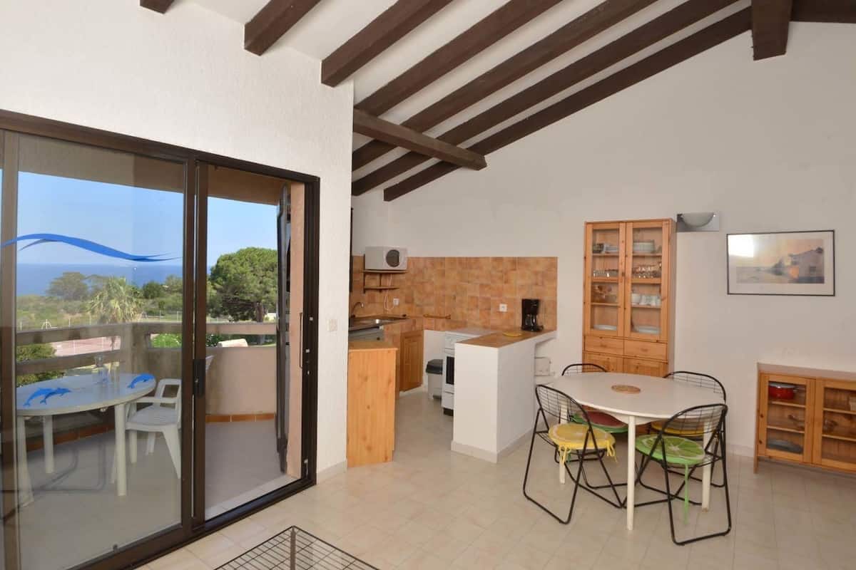 A modern kitchen with a view to the sea, a dining area with a table and chairs, and a wooden cabinet with dishes.