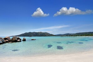 A beach with clear turquoise water, large rocks, and a distant mountain range under a blue sky.