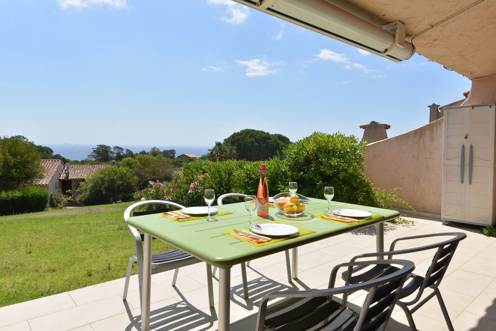 A dining table set outdoors with a view of the sea, surrounded by greenery and a building.