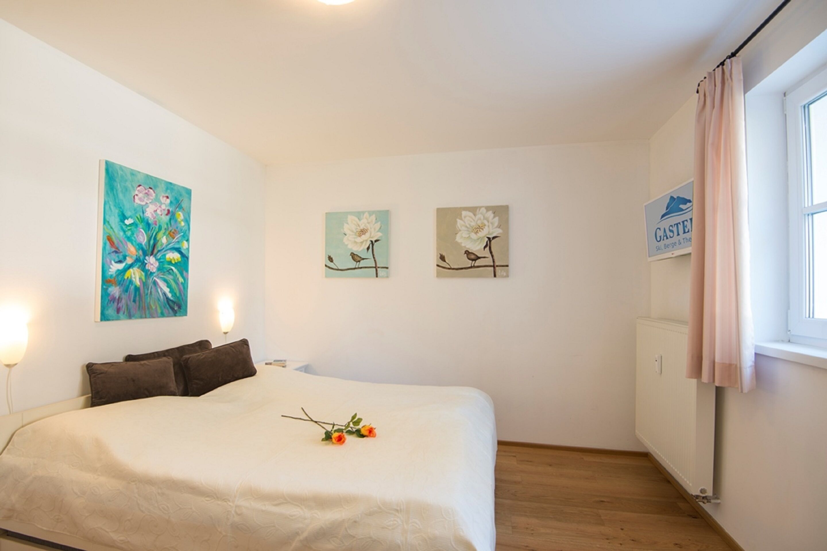 Ein Schlafzimmer mit einem Bett, zwei an der Wand befestigten Bildern, einem Heizkörper und einem Fenster mit Vorhängen.
