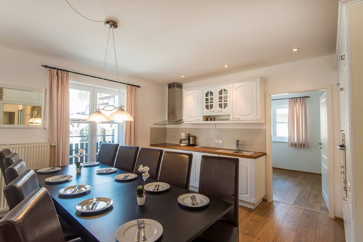 A modern dining room with a long table set for six, a kitchen with white cabinets, and a view through a large window.