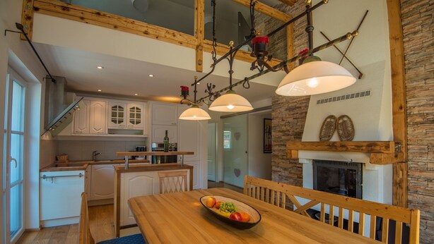 A modern kitchen with white cabinets, a wooden dining table with a bowl of fruit, and a stone fireplace. - Haus Wasmer by Alpentravel (Bad Hofgastein)