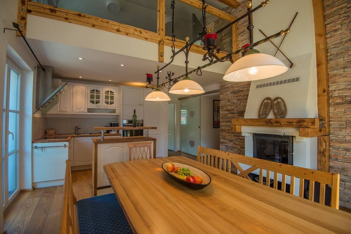 A modern kitchen with white cabinets, a wooden dining table with a bowl of fruit, and a stone fireplace.