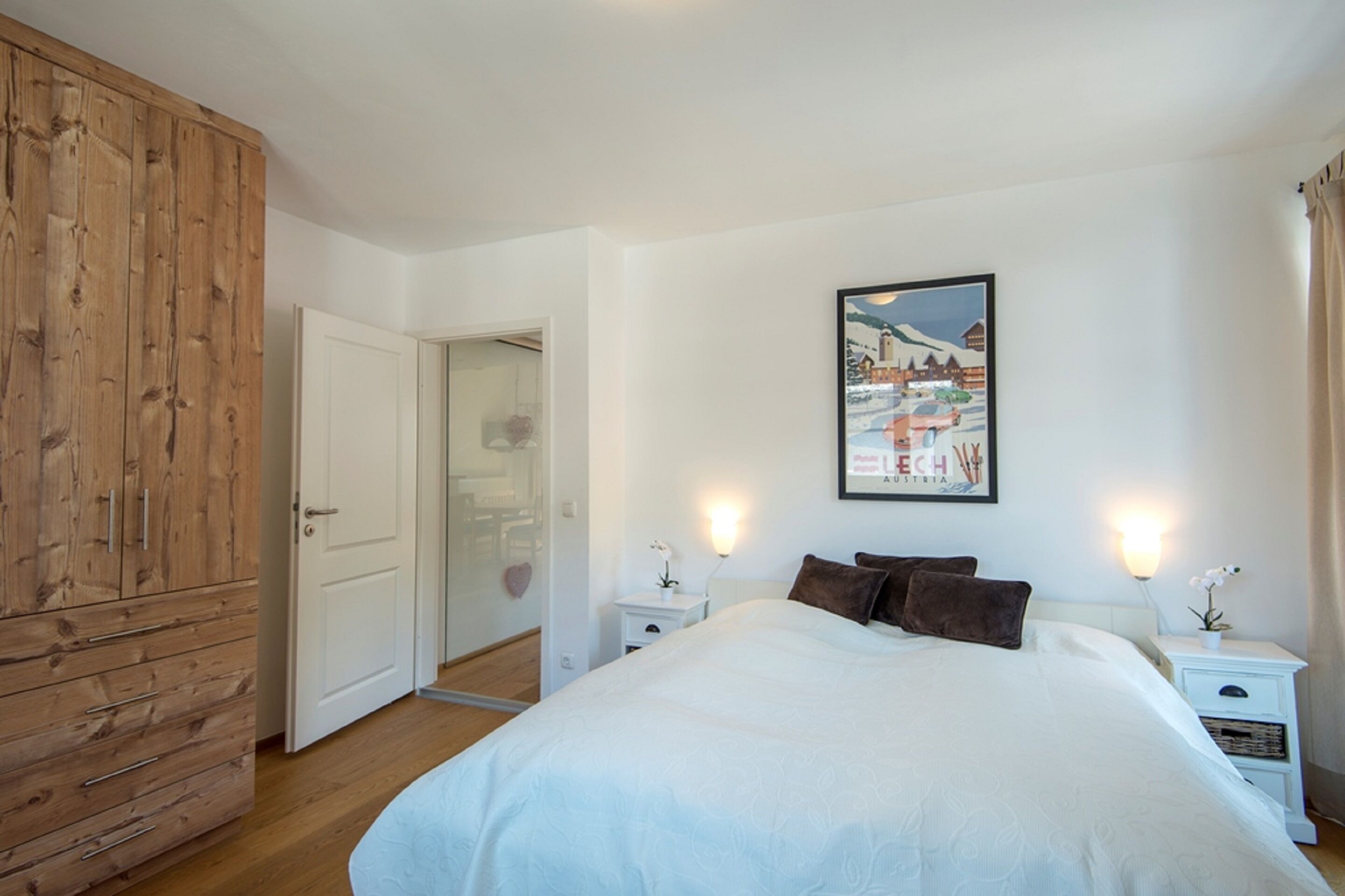A bedroom with a large bed, wooden wardrobe, white nightstands, and a framed picture on the wall.