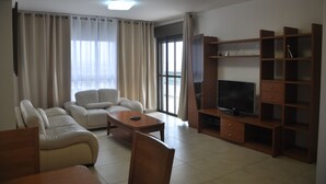 A living room with a sofa set, a coffee table, a TV on a wooden stand, and a large window with curtains.