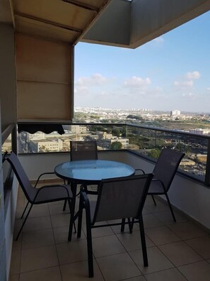 Varanda com mesa de vidro redonda e quatro cadeiras, com vista para um horizonte urbano com edifícios e vegetação.