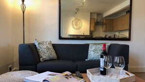Ett vardagsrum med en mörk soffa, mönstrade kuddar, ett soffbord med en tidning, ett brickbord med en flaska och glas, och en stor spegel som reflekterar ett kök.