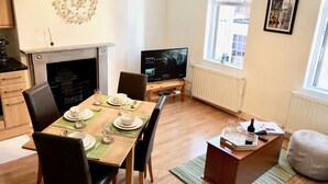 En matplats med ett träbord dukat för två personer, en öppen spis, en tv och ett fönster med utsikt över gatan.
