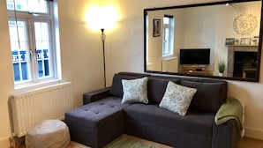Ett vardagsrum med en mörkgrå soffa, två mönstrade kuddar, en rund pall, en grön matta, en golvlampa, en stor spegel och en tv-apparat som står på ett träställ.