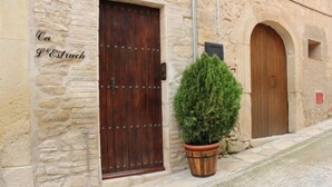A stone building with two wooden doors, one with metal studs, and a potted evergreen shrub in front. - Ca L'Estruch (Vallclara)
