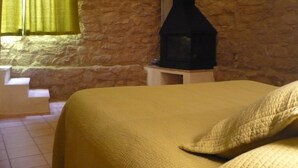 A room with a stone wall, a bed with a yellow bedspread, and a fireplace. - Ca L'Estruch (Vallclara)