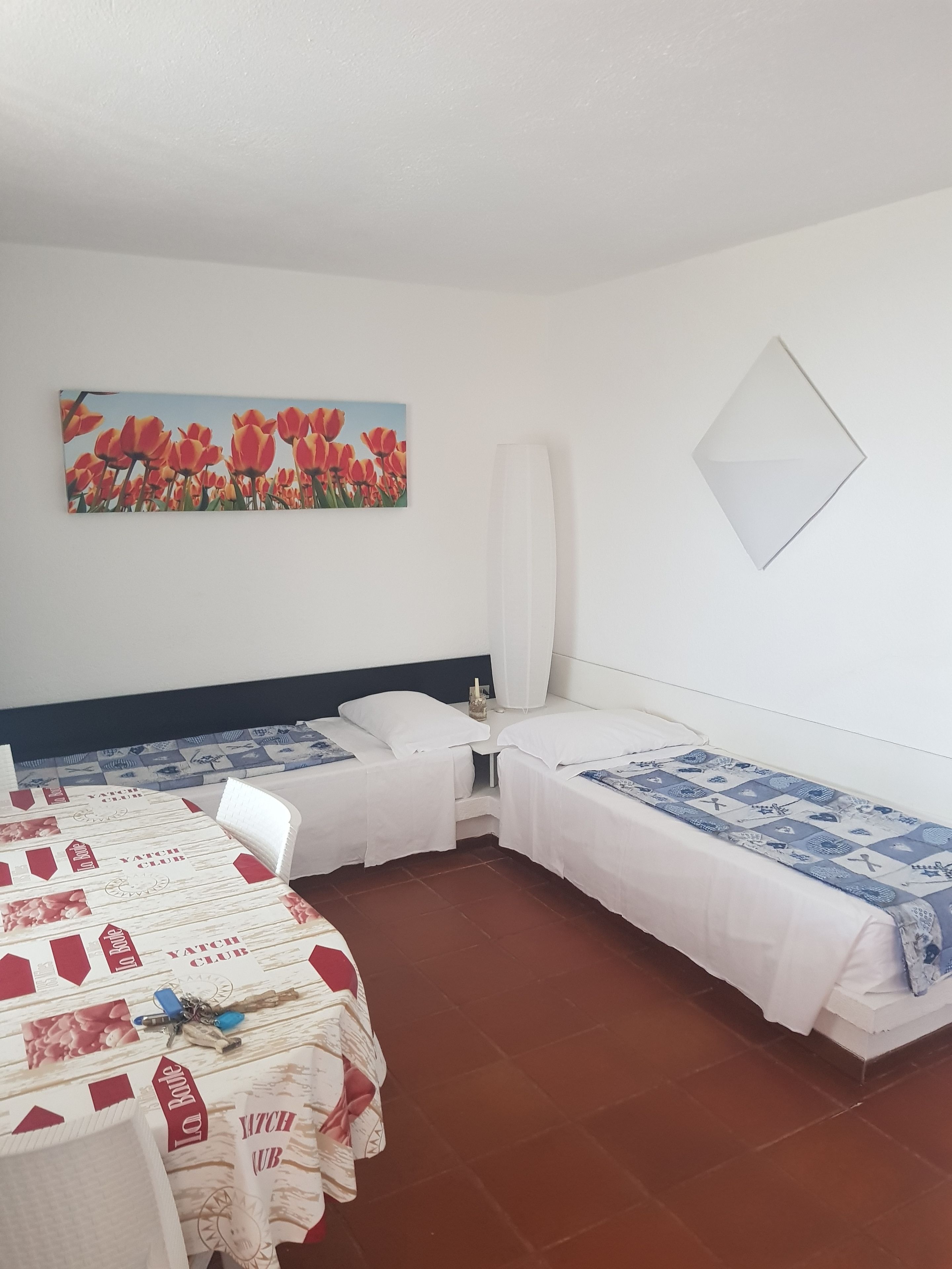 Una camera d'albergo con due letti singoli, un piccolo tavolo con tovaglia 'La Route', un dipinto di tulipani e uno specchio a parete.