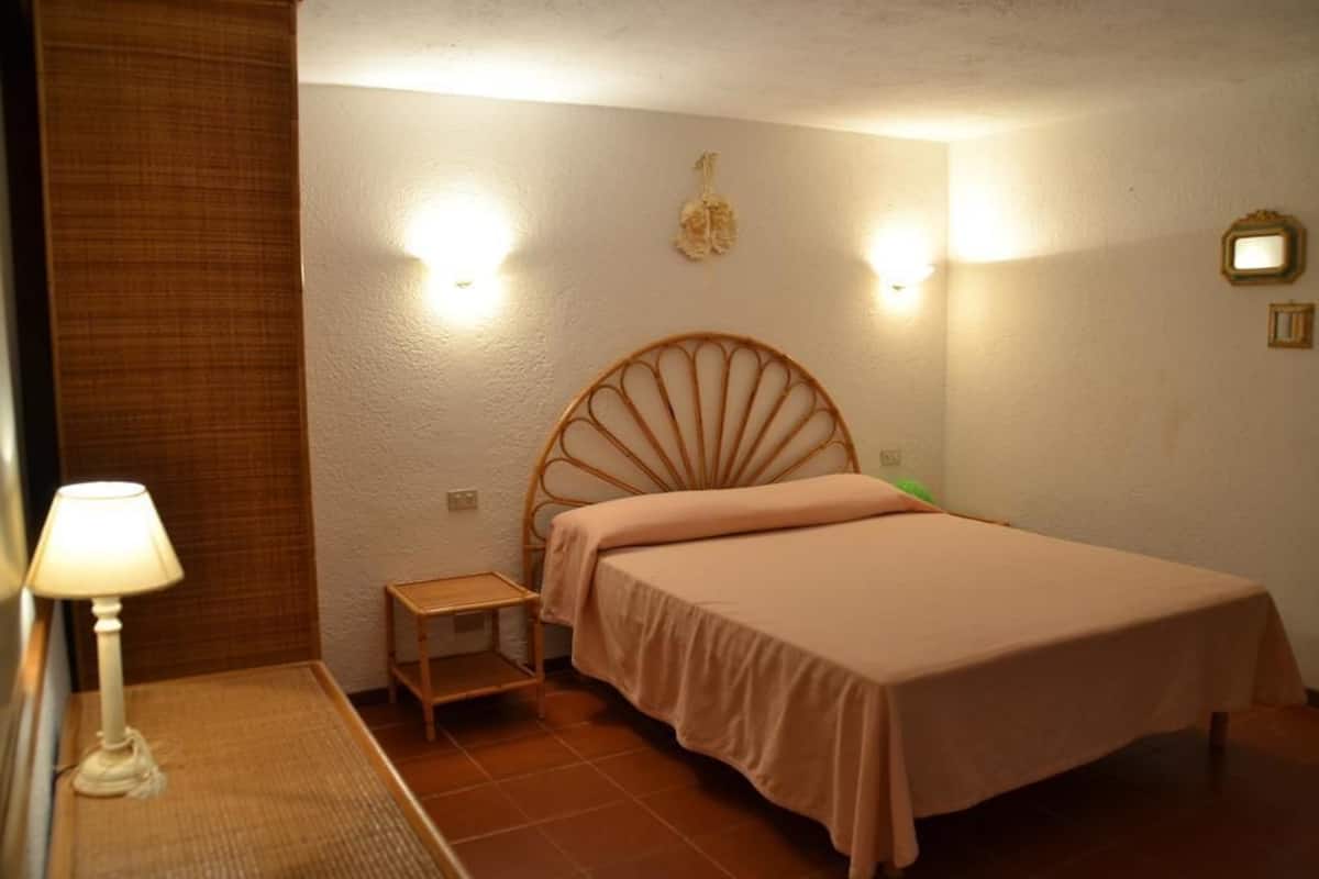 Una cama individual con cabecera de madera en una habitación de paredes blancas y suelo de baldosas. Hay una mesita de noche pequeña con una lámpara encima.