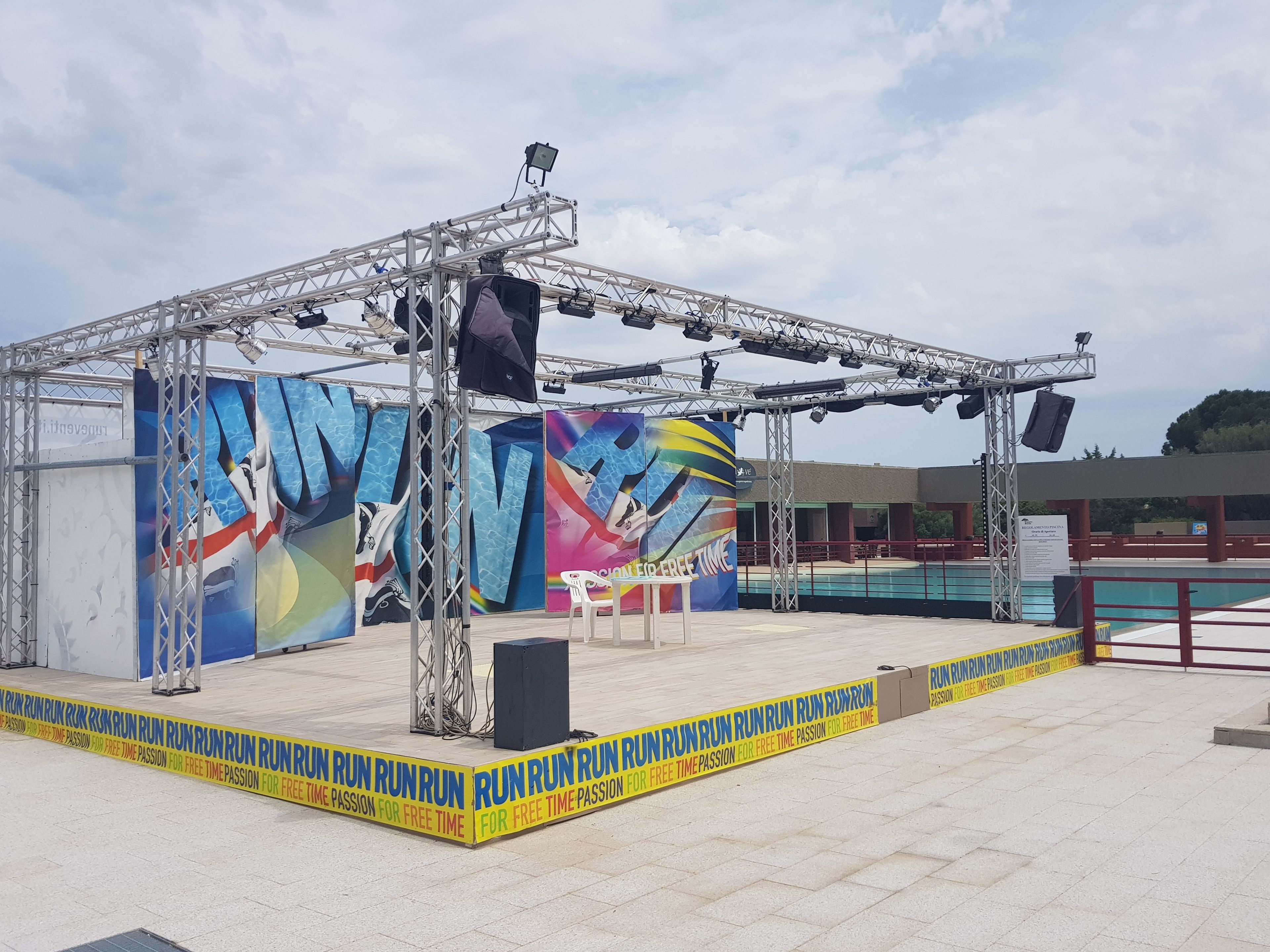 An outdoor stage with a metal frame, a large banner, and a pool in the background.