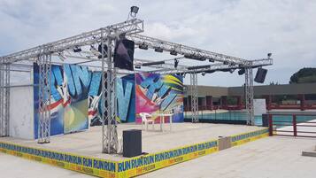 An outdoor stage with a metal frame, a large banner, and a pool in the background.