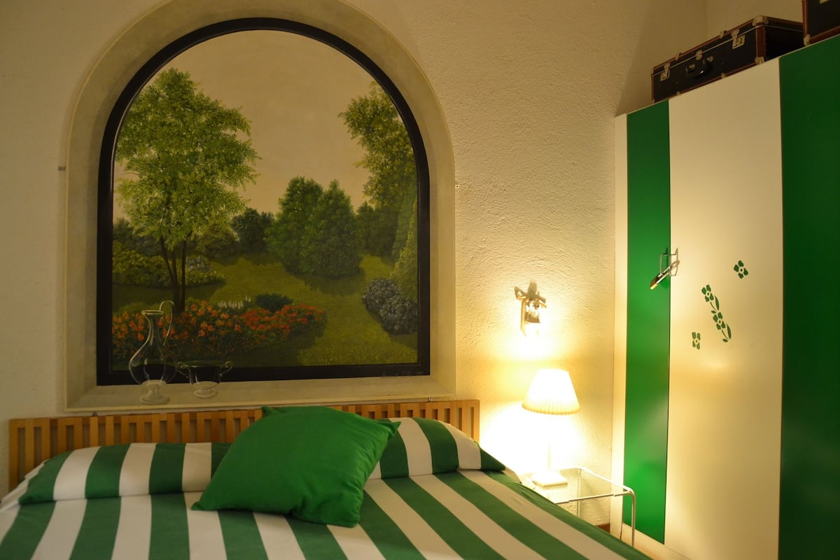 Una habitación con una cama con ropa de cama verde y blanca a rayas, un amplio ventanal con un cuadro de paisaje, una lámpara de noche y un armario verde.