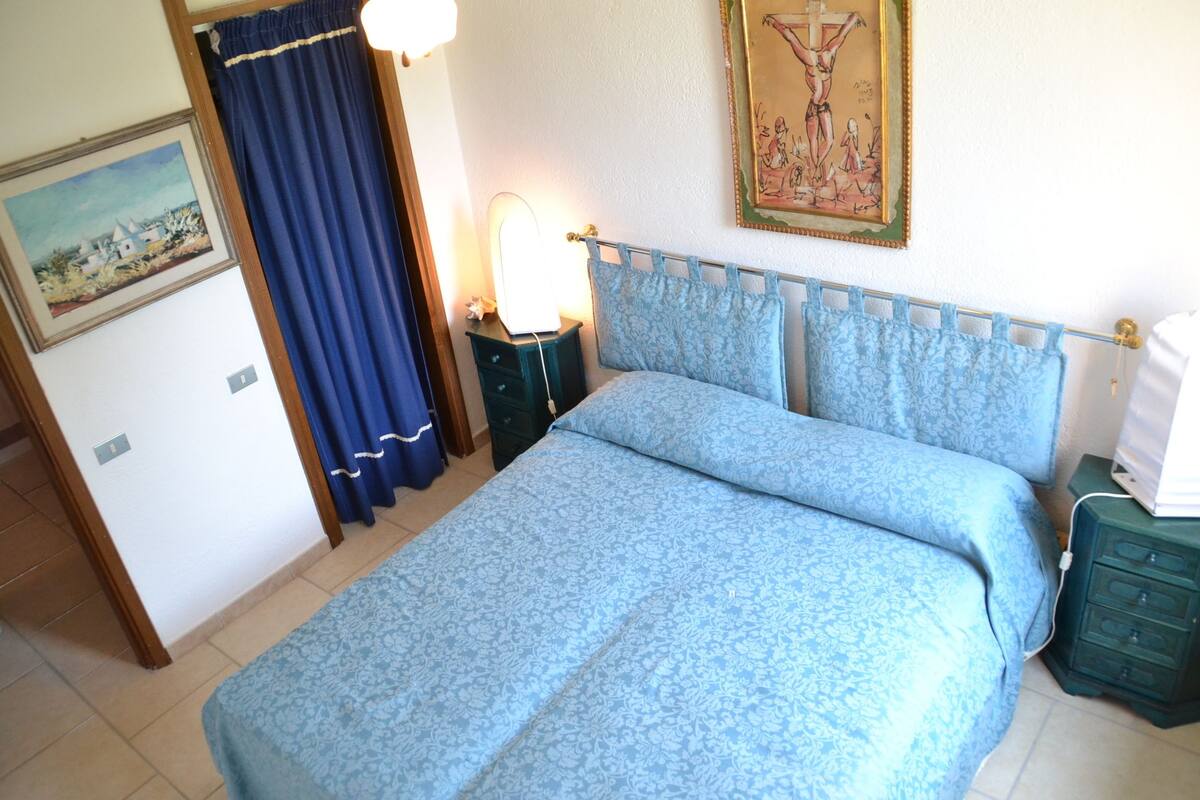 Un dormitorio con cama, mesita de noche, lámpara, cómoda, cuadro y cortina.