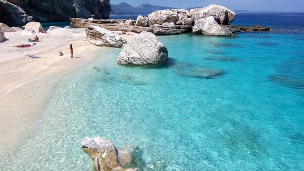Una playa con agua turquesa cristalina, grandes rocas y una orilla arenosa.