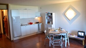 Una cocina compacta con armarios blancos, una mesa de comedor con sillas, un televisor pequeño y una lámpara de pared.
