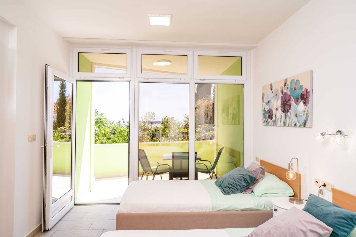 A bedroom with a bed, a nightstand, a lamp, a painting, and a balcony with chairs and a table.