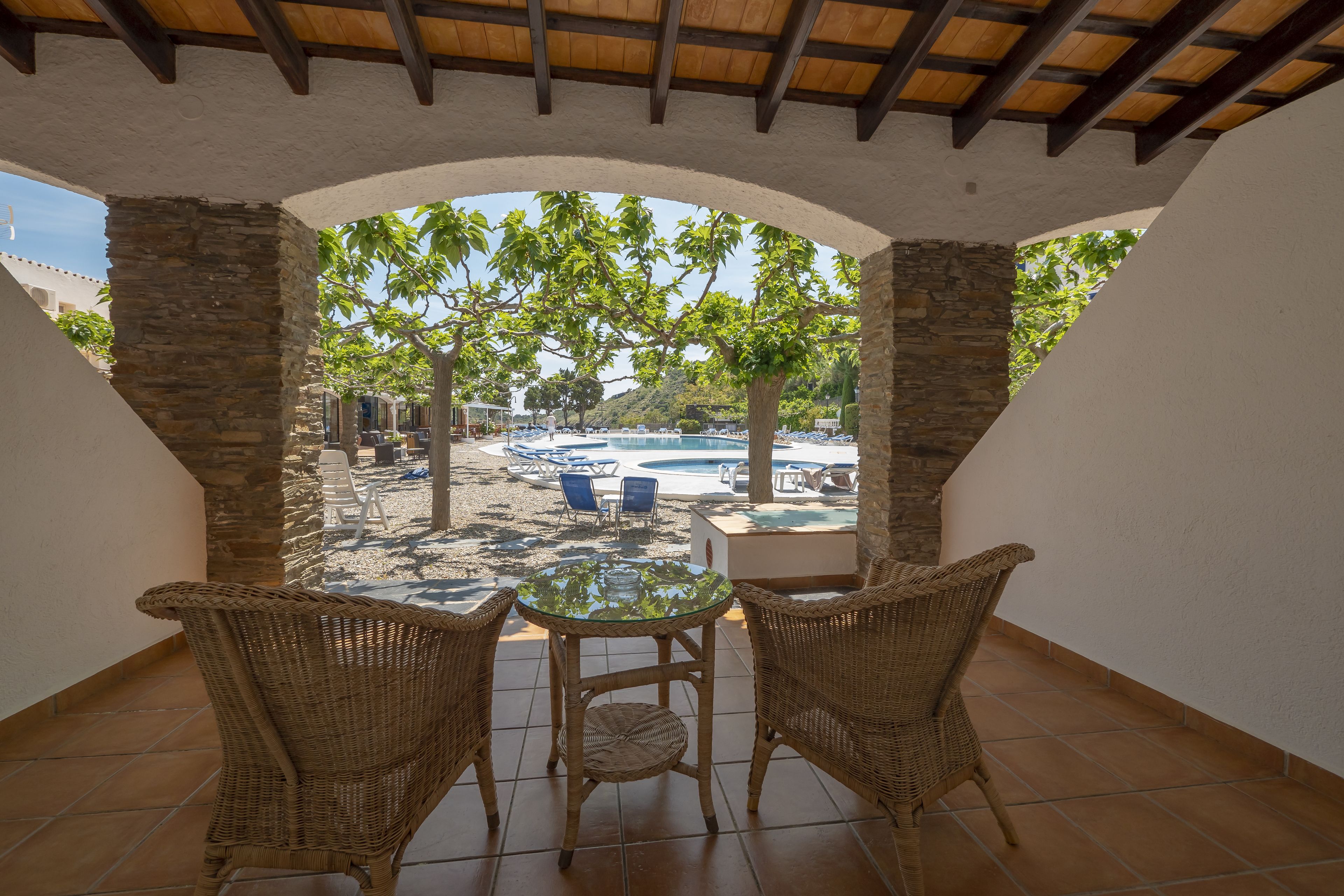 An outdoor patio with a glass table and wicker chairs, overlooking a pool area with lounge chairs and umbrellas.
