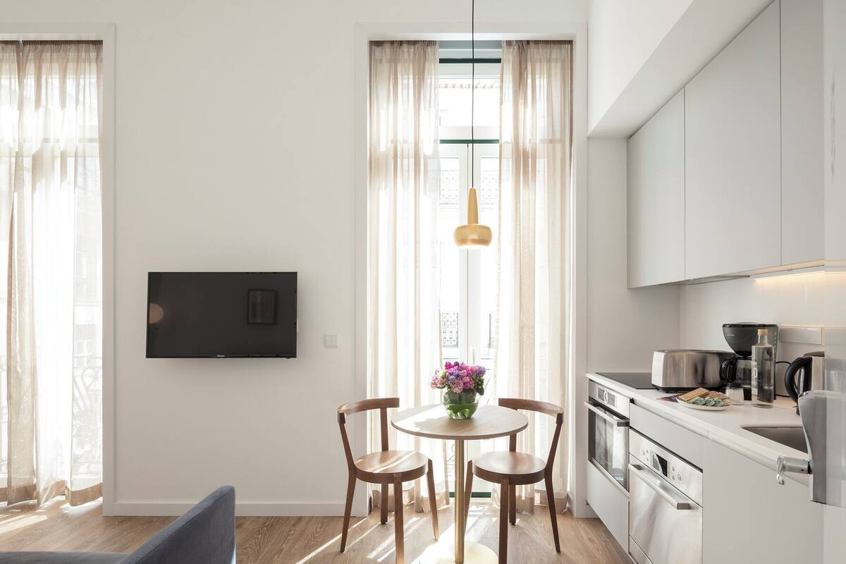A modern kitchen with white cabinets, a dining area with a round table and chairs, and a large window with sheer curtains.