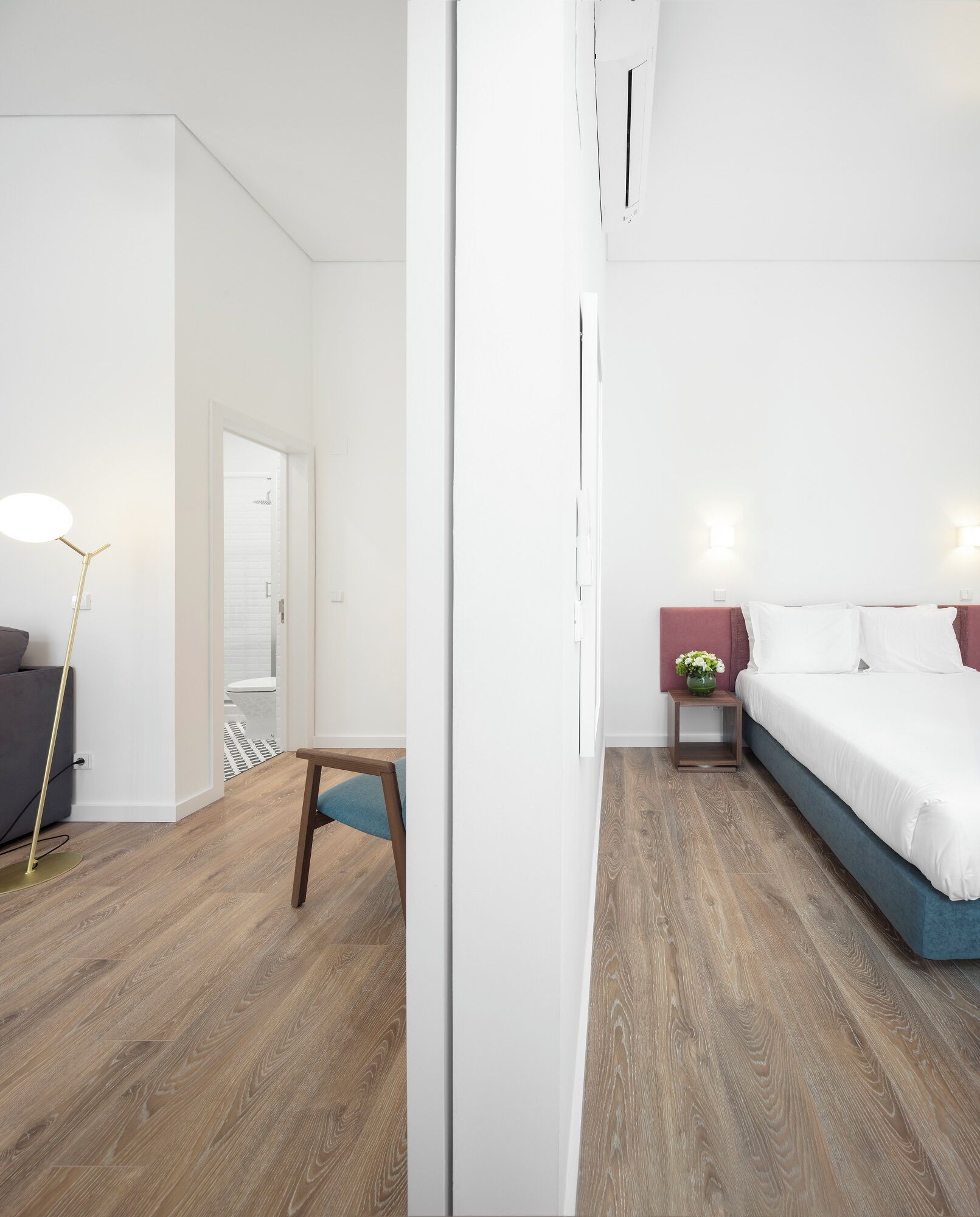 Moderni hotellihuone, jossa on puulattia, valkoisilla lakanoiden peitetty sänky, pieni pöytä tuolilla sekä lattiavalaisin.