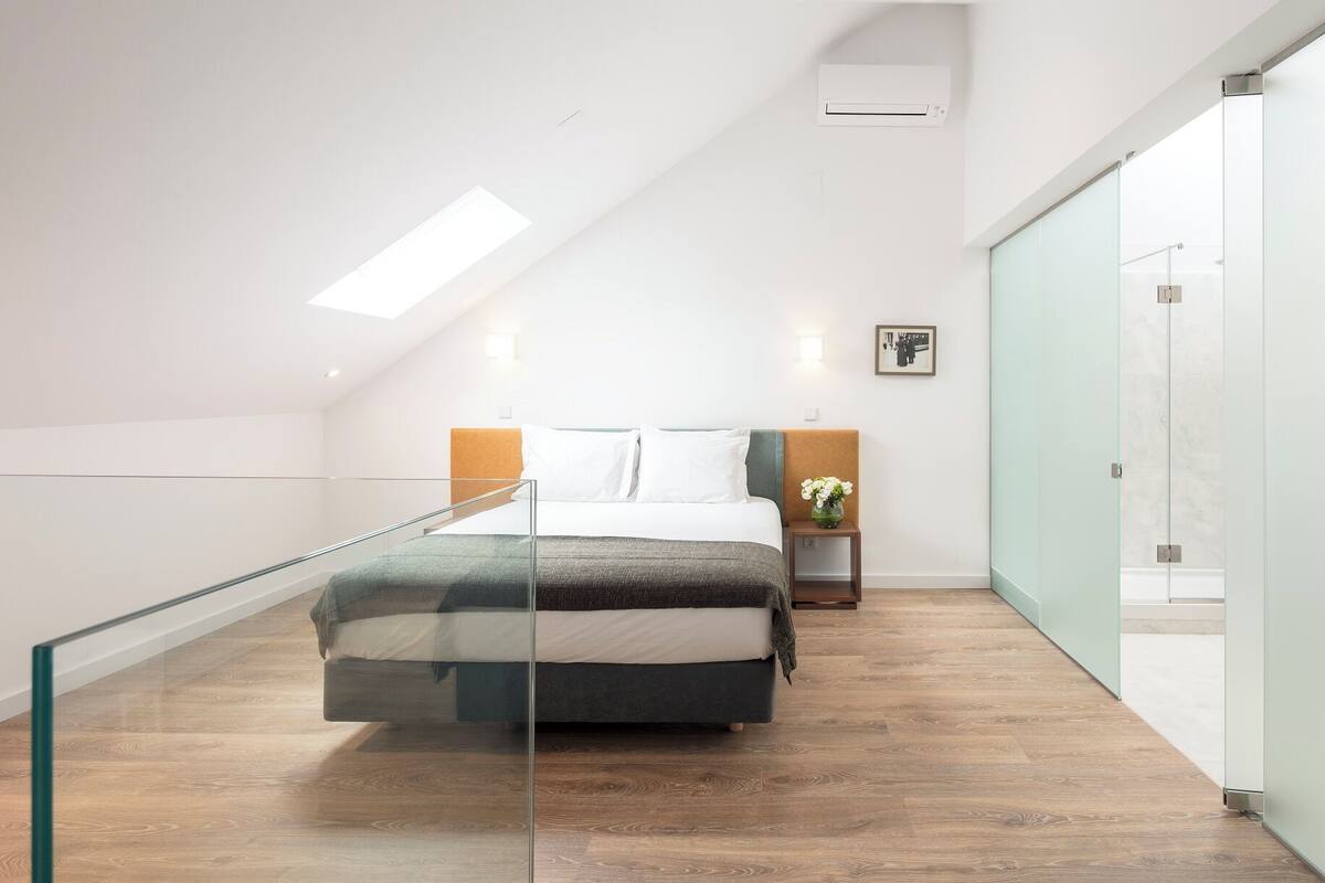 A modern bedroom with a glass railing, a bed with a headboard, a bedside table with a vase, and a wall-mounted air conditioner.