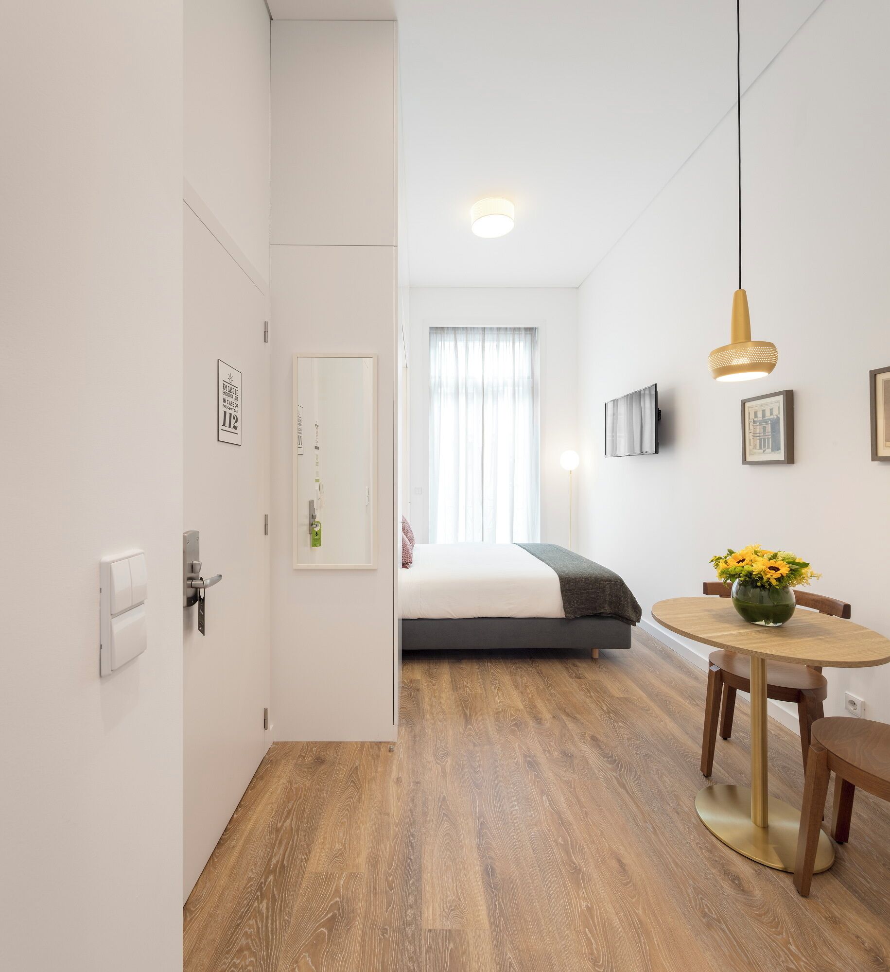 Quarto de hotel moderno com piso de madeira, cama com cabeceira cinza, mesa de jantar redonda com um vaso de flores e TV fixada na parede.