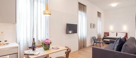 A modern living room with a sofa, a dining area with a table and chairs, a kitchen with a stove, and a TV mounted on the wall.