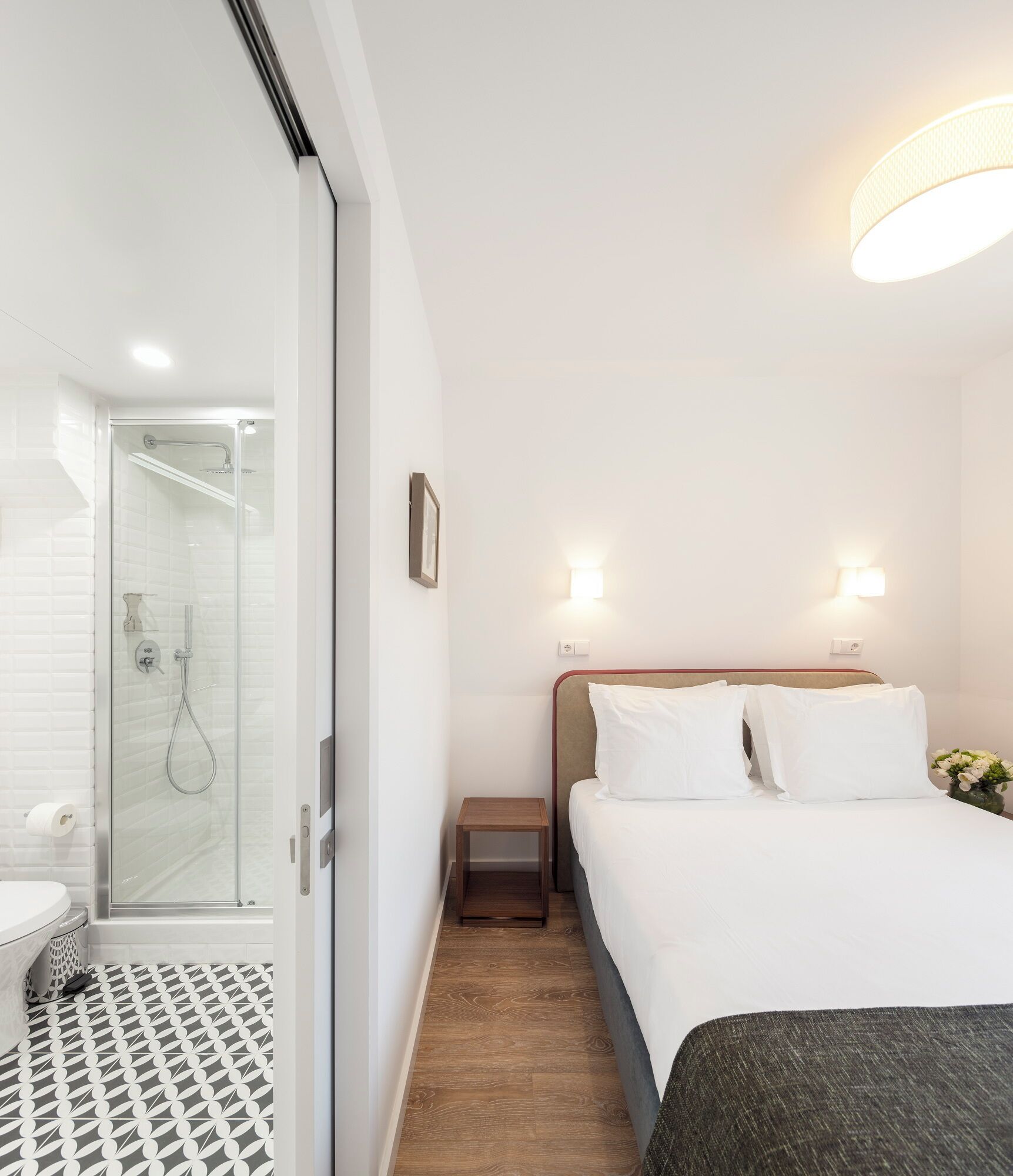 Hotellihuone, jossa on suihku, valkoisilla lakanoiden peitetty sänky, yöpöytä ja seinään kiinnitetty valaisin.