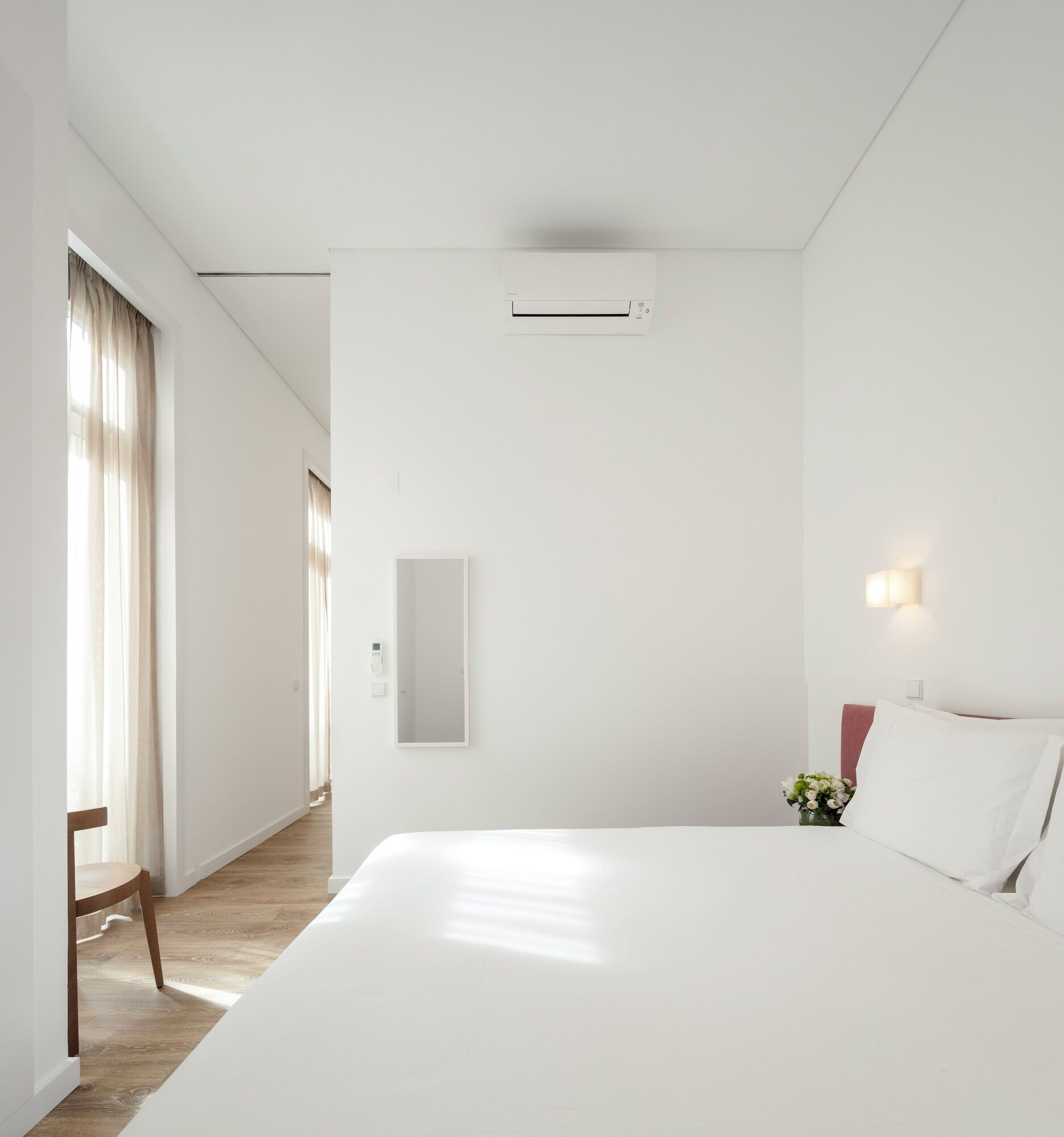 A hotel room with a large bed, a bedside table with a vase of flowers, a wall-mounted air conditioning unit, and a mirror.