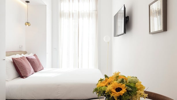A modern hotel room with a bed, a round wooden table with a vase of flowers, and a large window with sheer curtains.