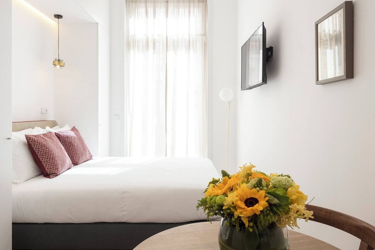 A modern hotel room with a bed, a round wooden table with a vase of flowers, and a large window with sheer curtains.